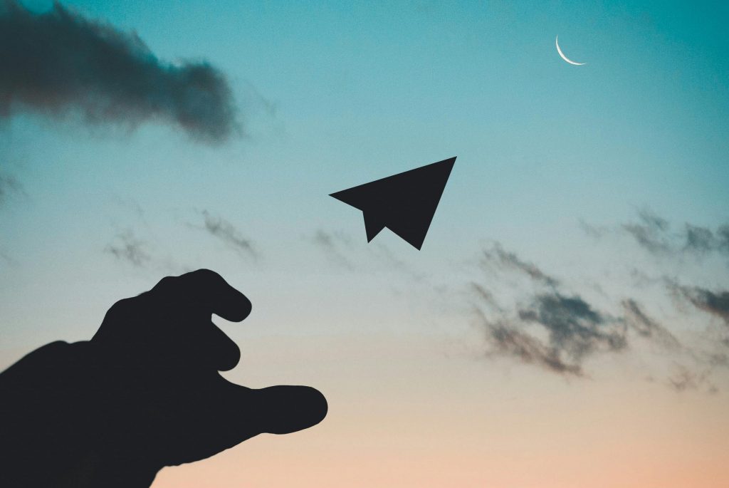 A silhouette of a hand launching a paper plane against a colorful dusk sky with a crescent moon.